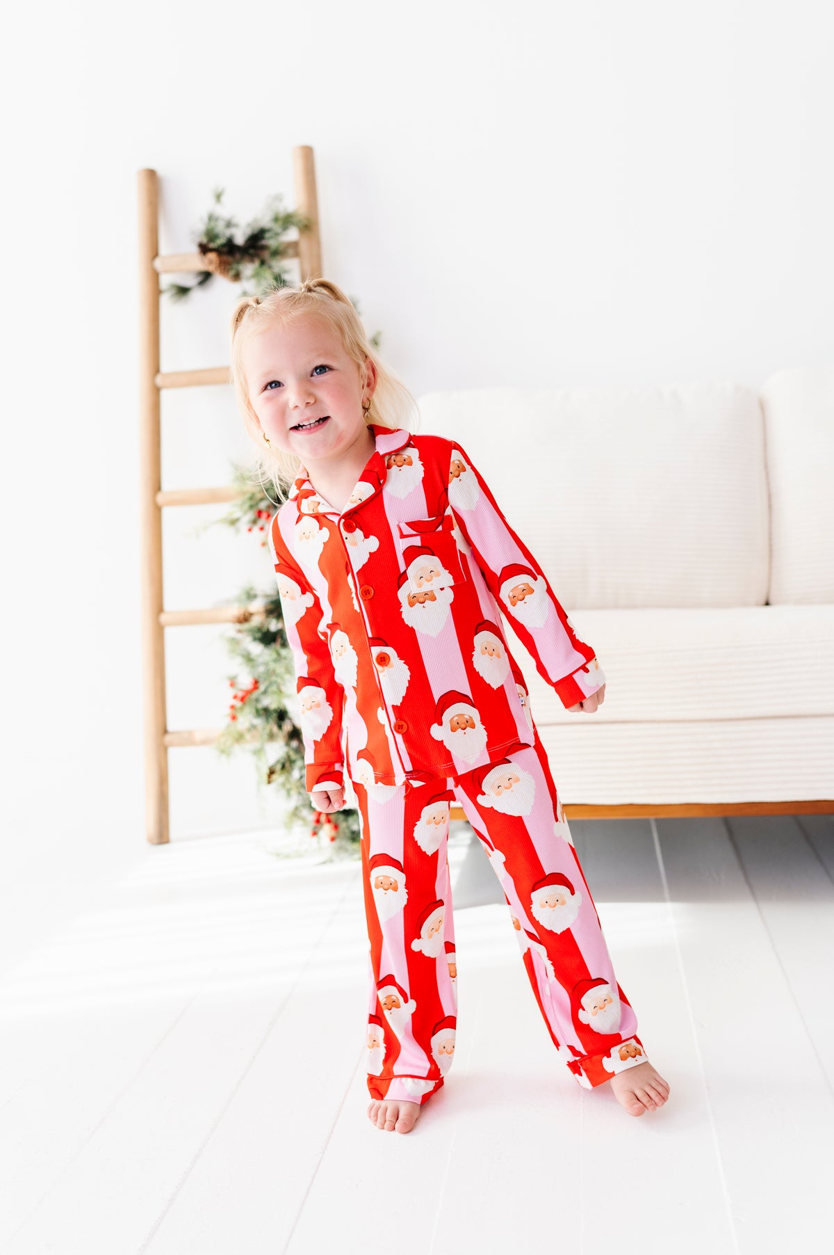 A young girl with blonde pigtails stands barefoot in a bright room, wearing a red and white Santa Claus patterned bamboo pajama set. A wooden ladder with greenery and a white sofa are visible in the background.