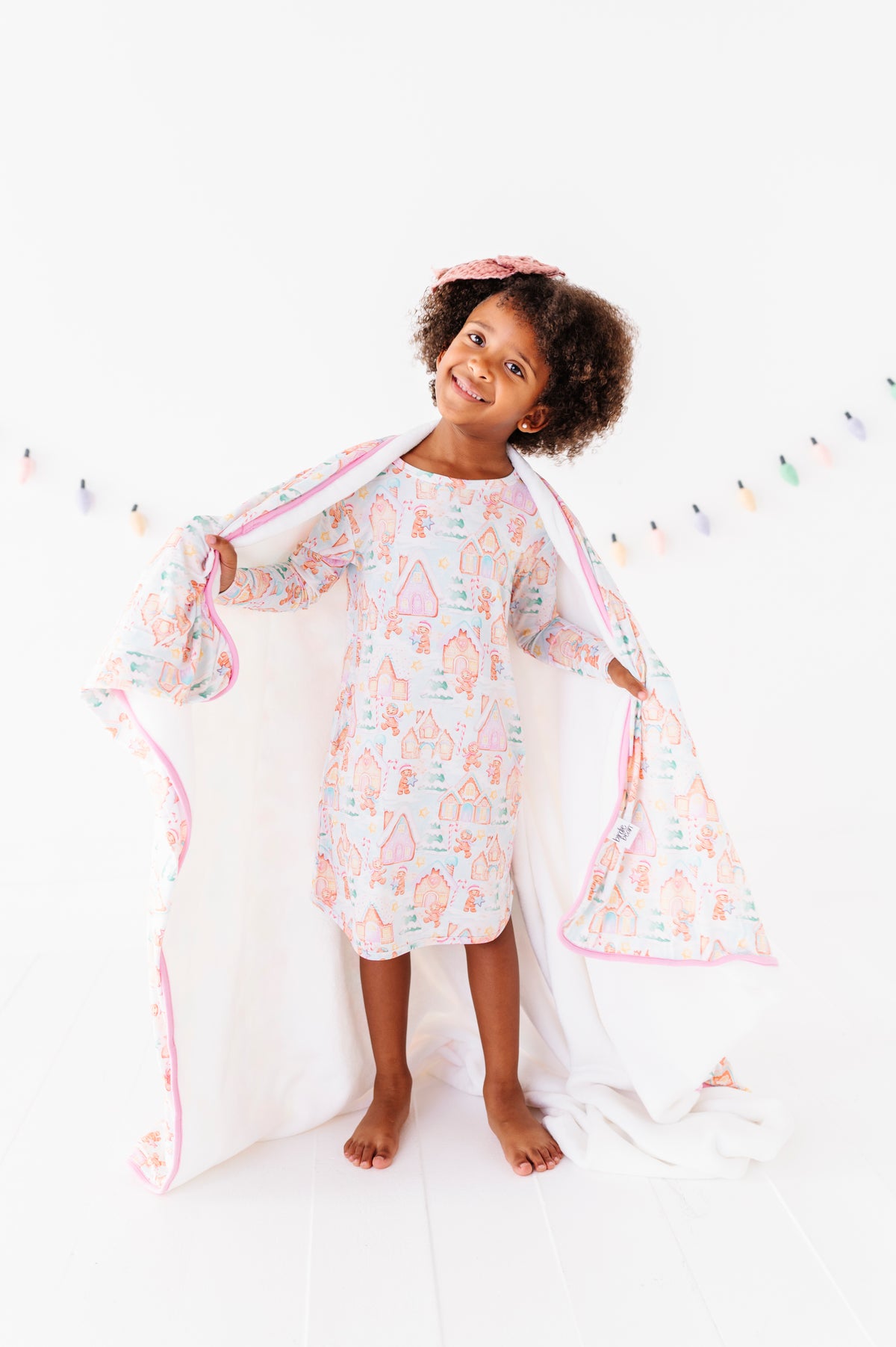 A little girl standing in a bright white room, wearing a Christmas gingerbread village themed bamboo gown and wrapped in a matching bamboo plush blanket 