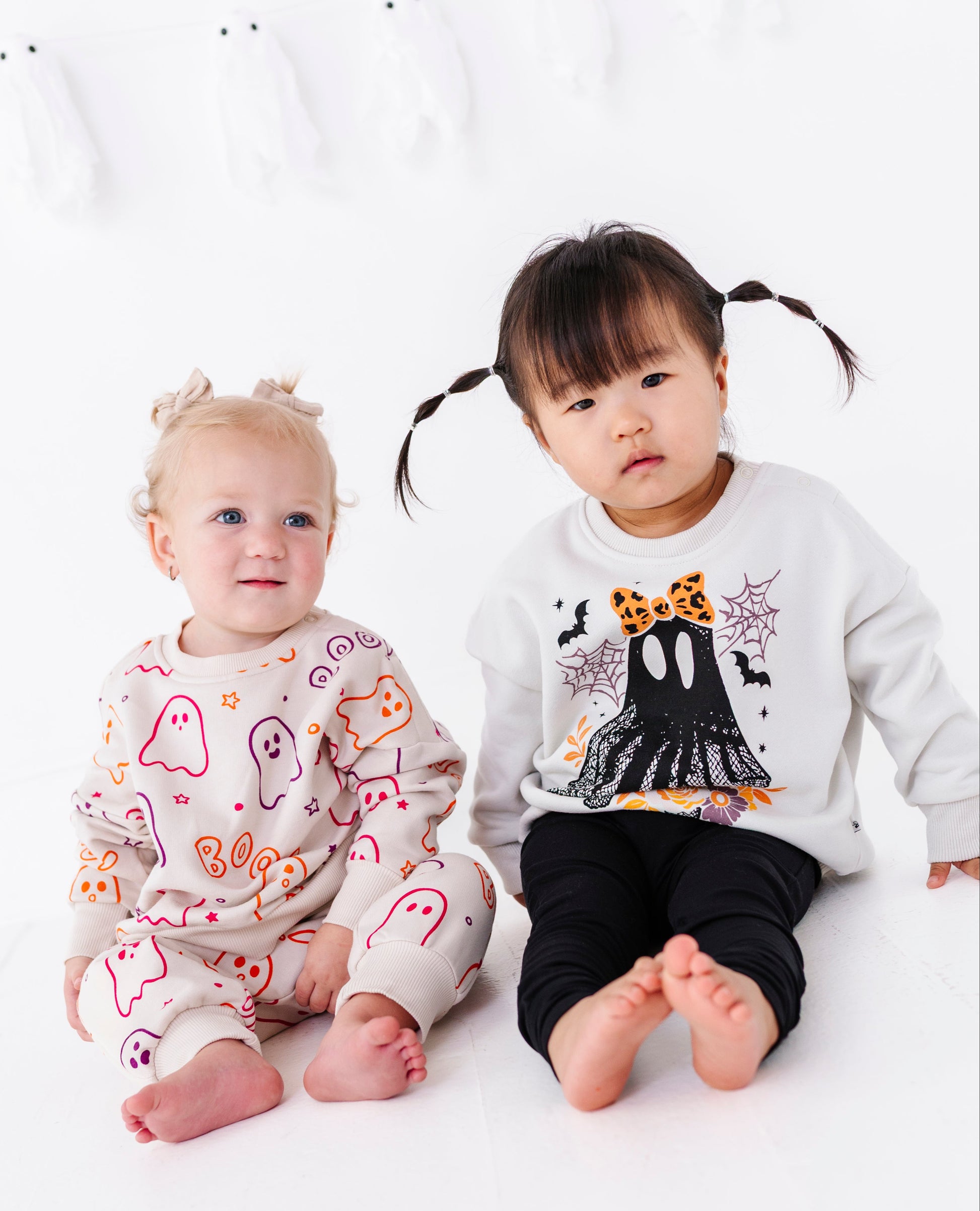 Two toddlers sitting on a white floor wearing matching Halloween-themed outfits. The toddler on the right wears a white long-sleeve top with a black ghost graphic and black pants, while the toddler on the left wears a light beige jogger set with colorful ghost patterns.