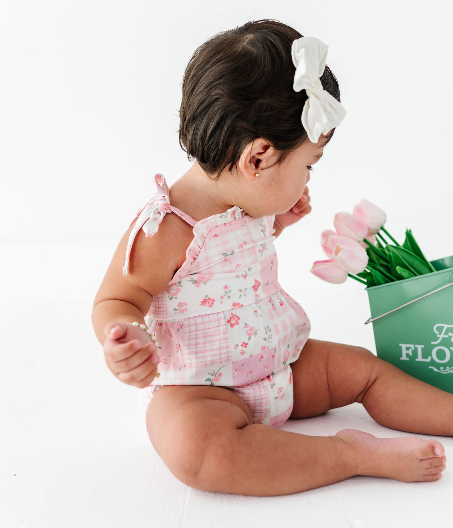 Baby girl in a floral bamboo outfit holding pink tulips next to a green bucket with flowers on a white background