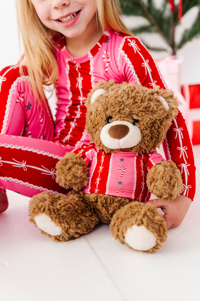 A young girl in festive red and pink bamboo pajamas sits on the floor smiling while holding a brown plush teddy bear wearing a matching red and pink holiday tee.