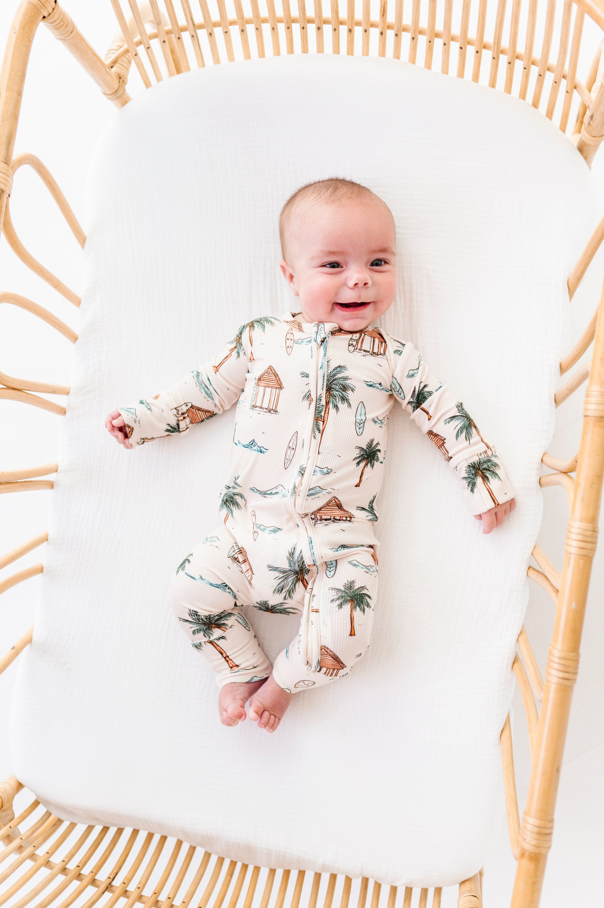 Baby in a beach patterned onesie lying in a rattan crib with white mattress