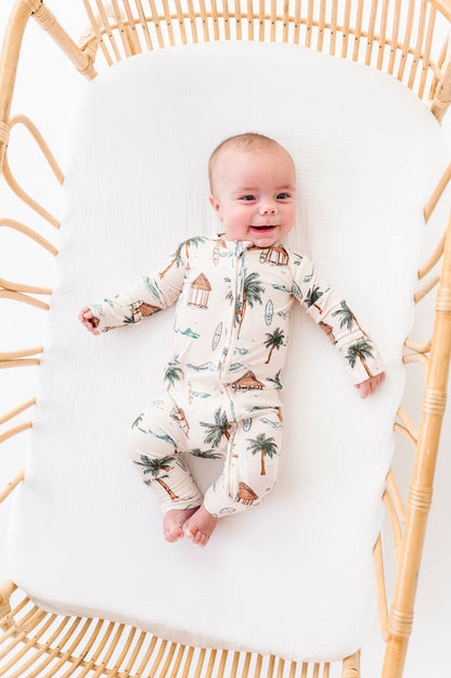 Baby in a beach patterned onesie lying in a rattan crib with white mattress