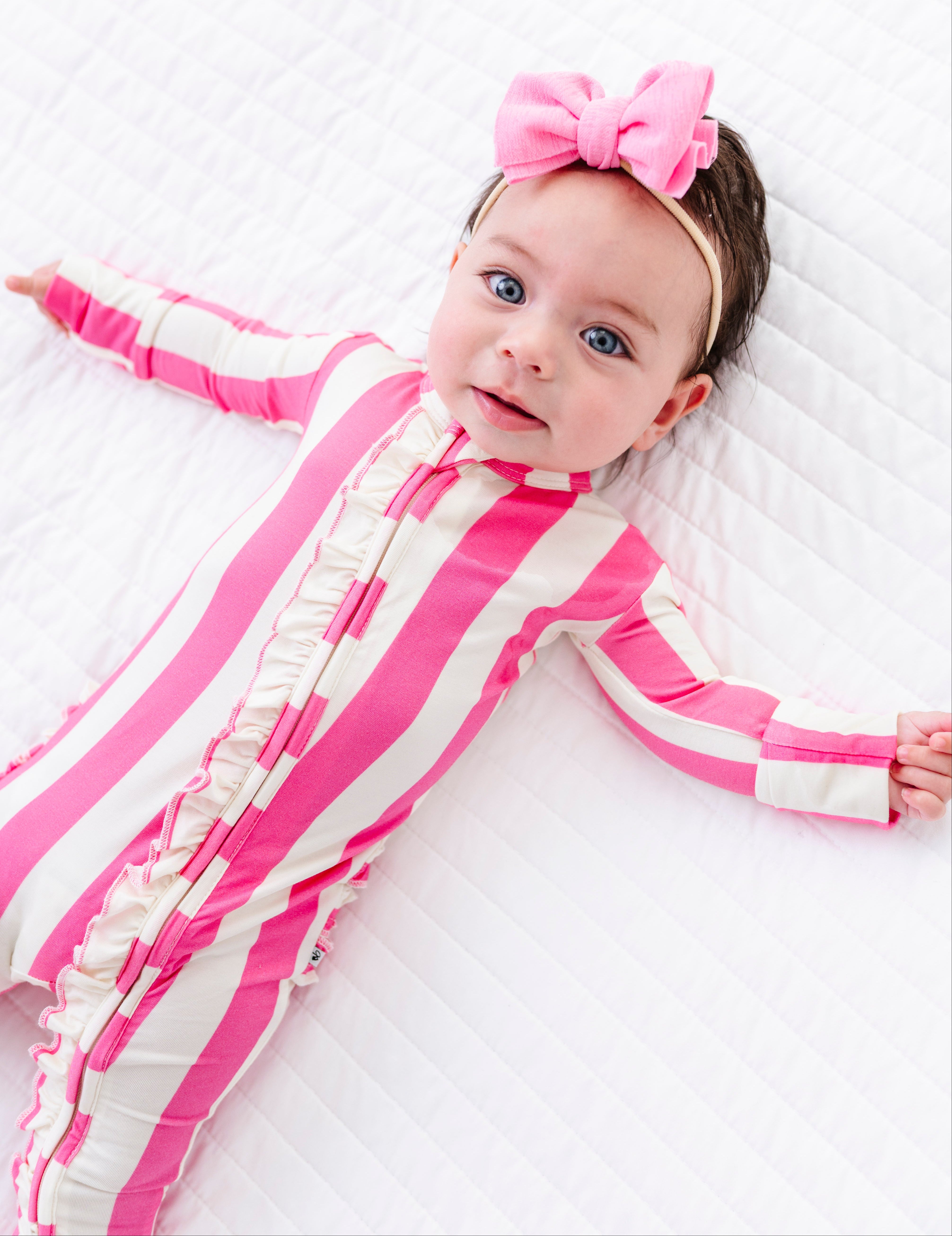A baby lying on a white quilted blanket, wearing a pink and white striped long-sleeve footless onesie with ruffle trim and a matching pink bow headband.