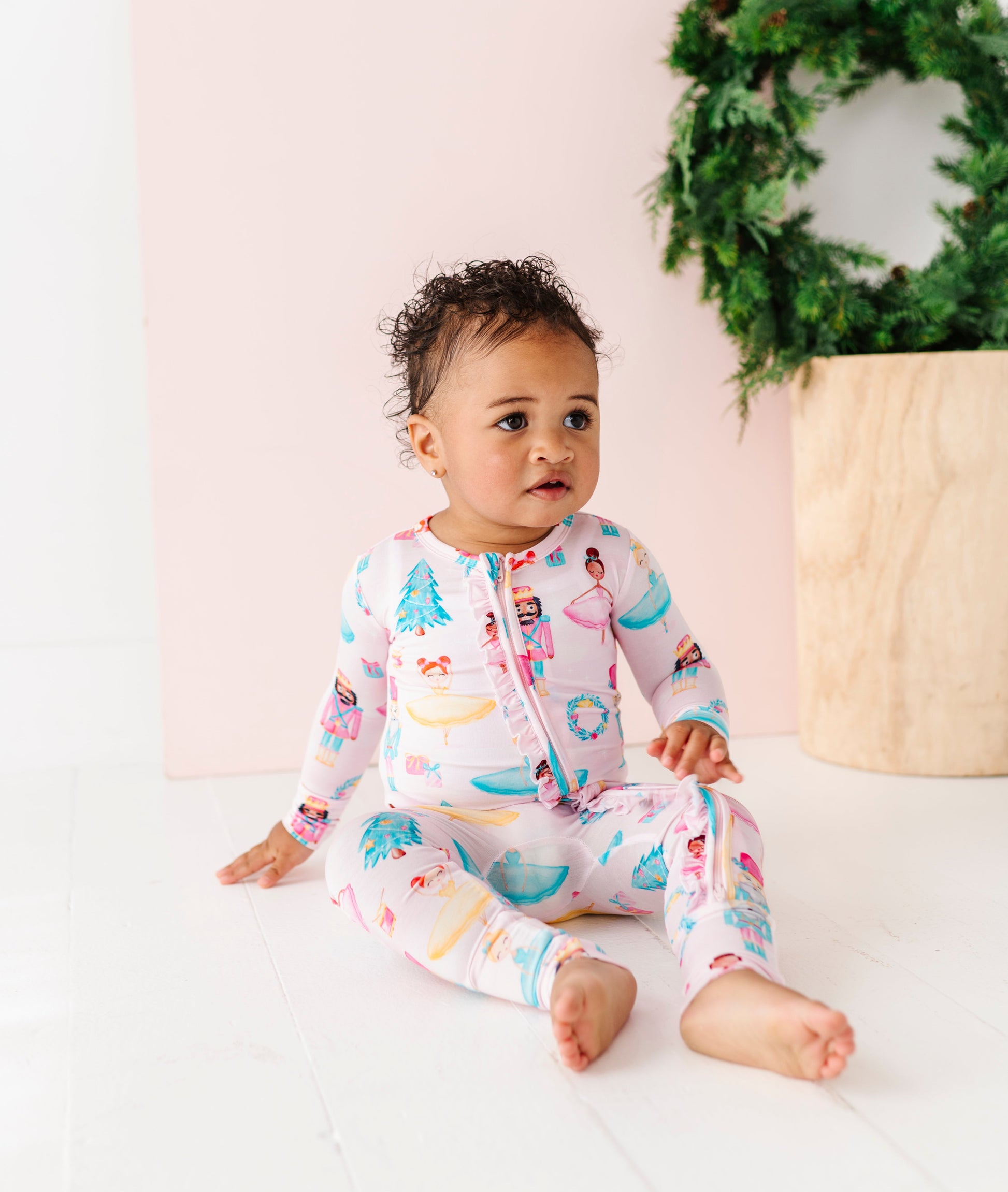 Baby in a Christmas bamboo zip romper sitting on a white surface with a decorative wreath in the background.