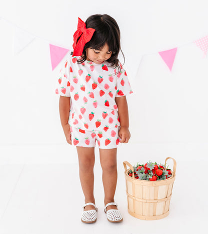 Child wearing a white outfit with strawberry patterns, standing next to a basket of strawberries on a white background.