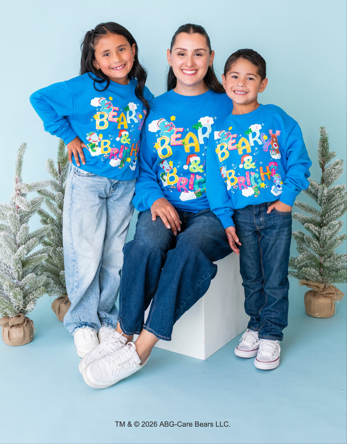 Three people posing together in matching bright blue long-sleeve shirts with a colorful 'BEAR & BRITISH' graphic, set against a light blue backdrop with small potted trees.