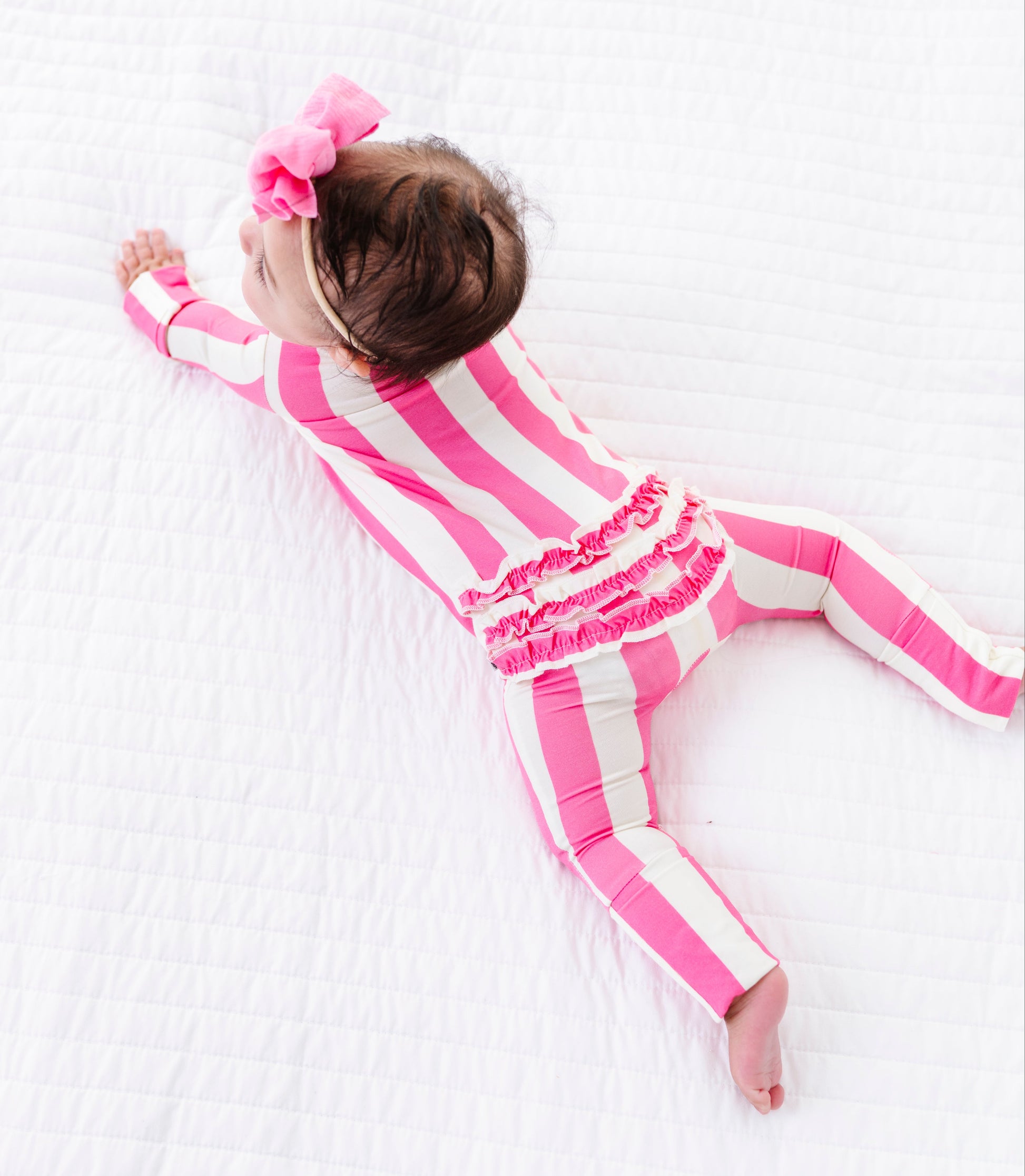 A baby lying on a white quilted blanket, wearing a pink and white striped long-sleeve romper with a ruffled waistband and a matching pink bow headband.