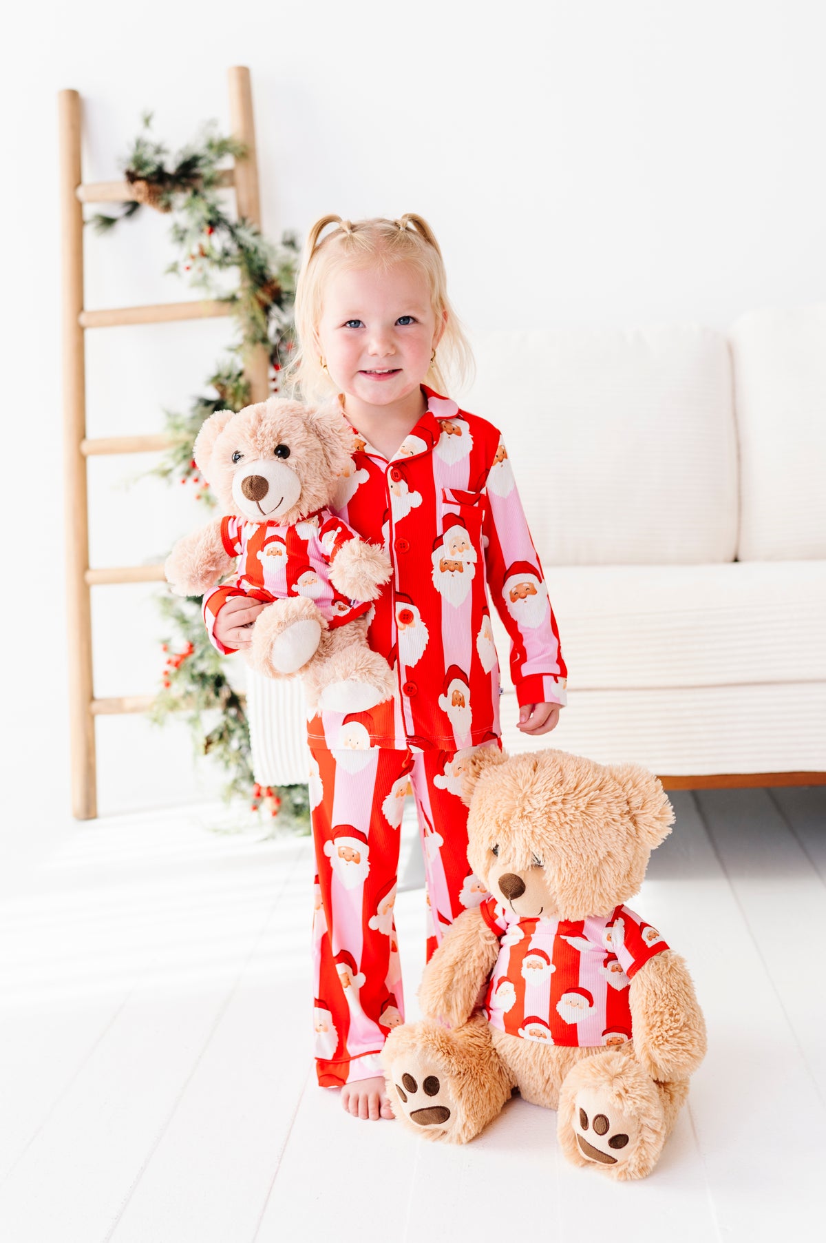 A young girl with blonde pigtails stands barefoot in a bright room wearing red Santa-patterned bamboo pajamas and holding a teddy bear. A larger teddy bear sits in front of her, also wearing a matching red Santa pajama set.