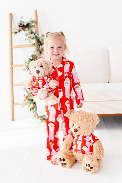 A young girl with blonde pigtails stands barefoot in a bright room wearing red Santa-patterned bamboo pajamas and holding a teddy bear. A larger teddy bear sits in front of her, also wearing a matching red Santa pajama set.
