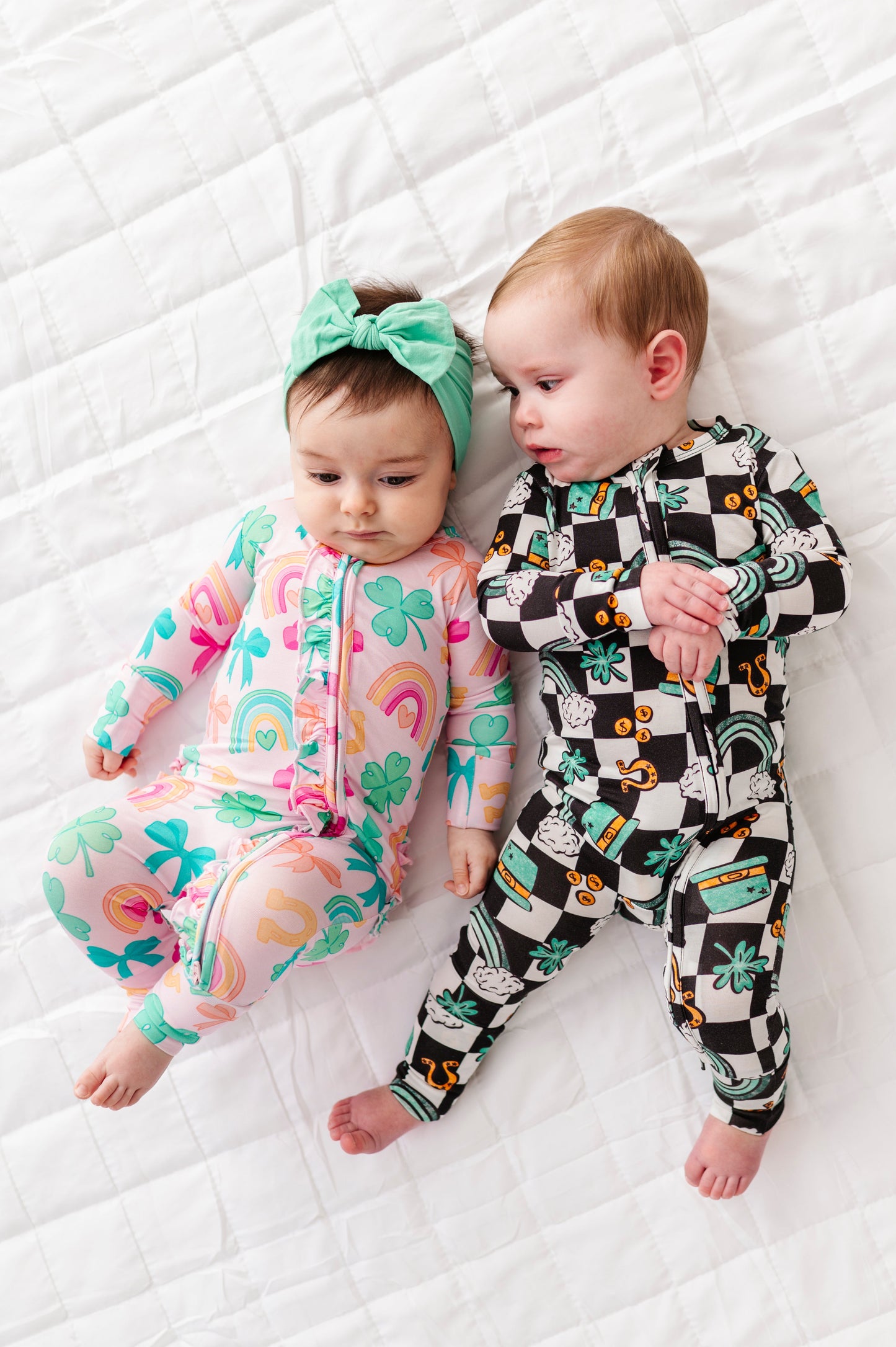 Two babies wearing colorful St. Patrick's Day themed bamboo onesies lying on a white textured blanket