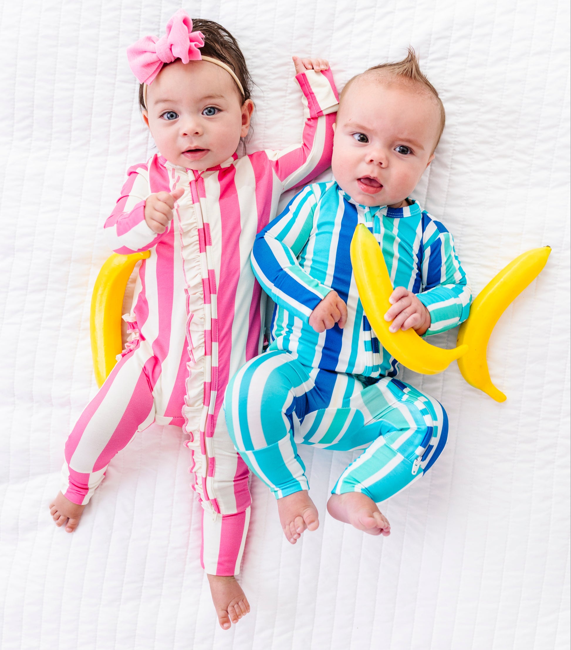 Two babies lying on a white textured blanket, wearing matching banana-themed onesies. The baby on the left wears a pink and white striped romper with a pink bow in her hair, and the baby on the right wears a blue and white striped romper while holding a yellow banana prop.