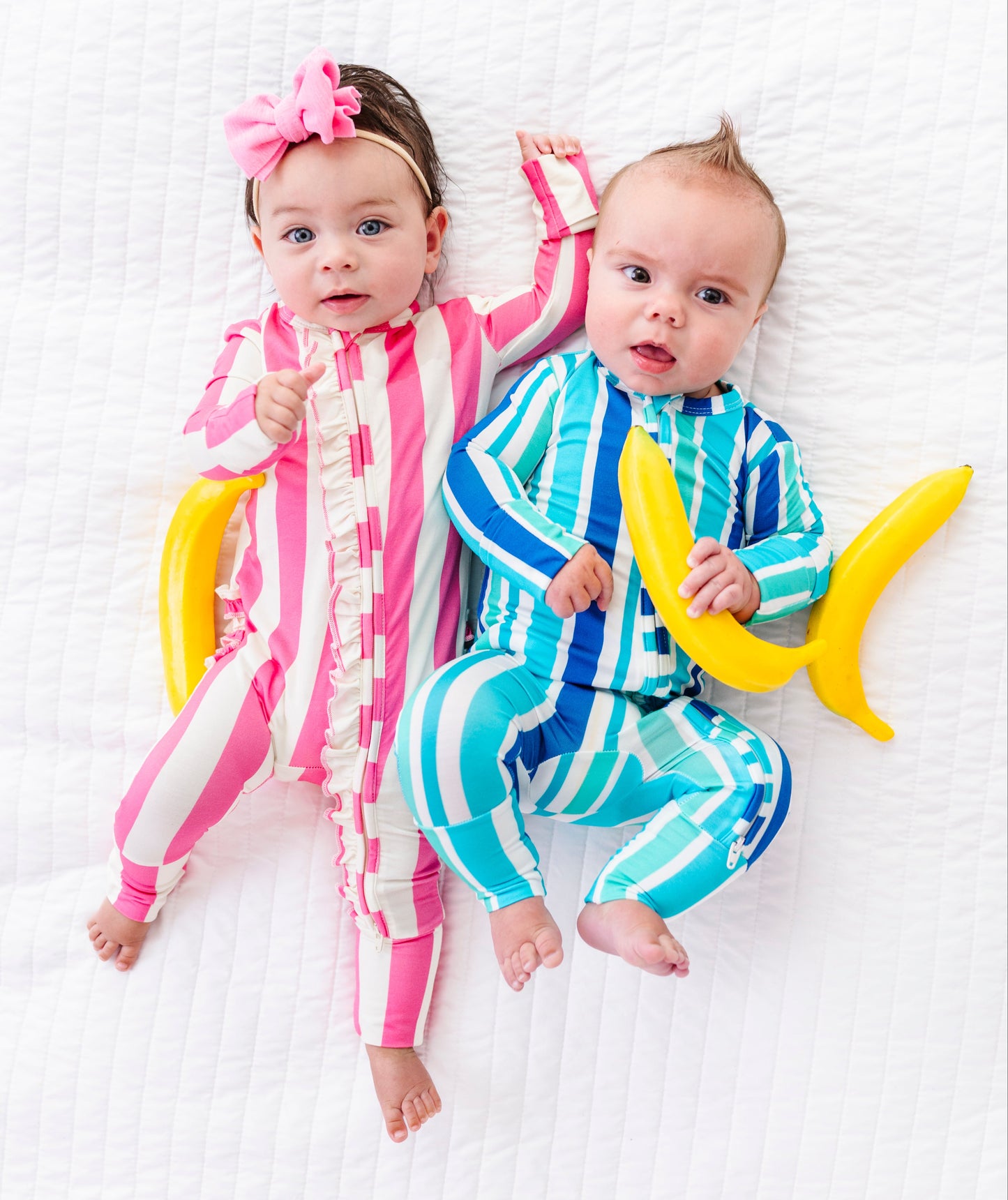 Two babies lying on a white textured blanket, wearing matching banana-themed onesies. The baby on the left wears a pink and white striped banana jumpsuit with a pink bow in her hair, and the baby on the right wears a blue and white striped banana jumpsuit while holding a yellow banana prop.