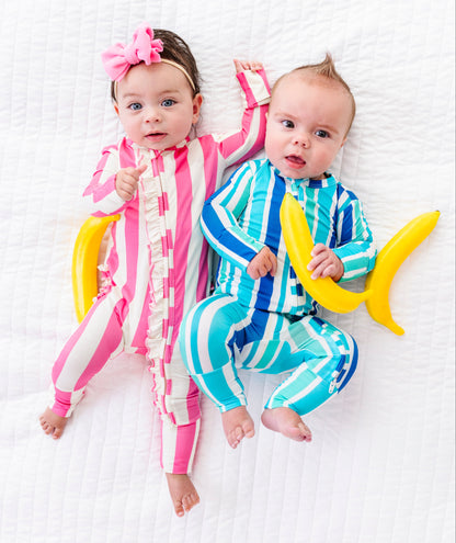 Two babies lying on a white textured blanket, wearing matching banana-themed onesies. The baby on the left wears a pink and white striped banana jumpsuit with a pink bow in her hair, and the baby on the right wears a blue and white striped banana jumpsuit while holding a yellow banana prop.