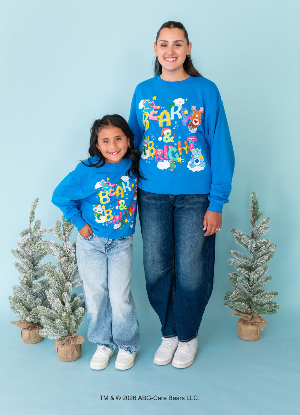A woman and a child wearing matching bright blue sweatshirts with a colorful 'Beach & Barbershop' graphic, standing against a light blue background with two small potted flocked trees.