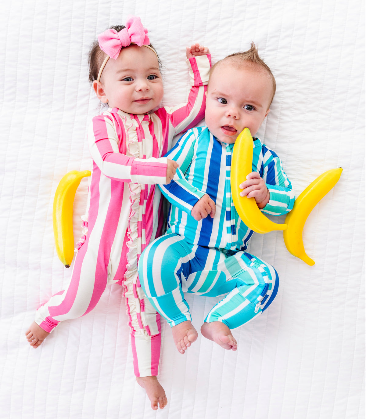 Two babies lying on a white textured blanket, wearing matching striped pajamas. The baby on the left wears pink and white vertical stripes with a pink bow headband, while the baby on the right wears blue and white vertical stripes and holds a yellow banana toy.