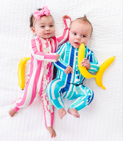 Two babies lying on a white textured blanket, wearing matching striped pajamas. The baby on the left wears pink and white vertical stripes with a pink bow headband, while the baby on the right wears blue and white vertical stripes and holds a yellow banana toy.