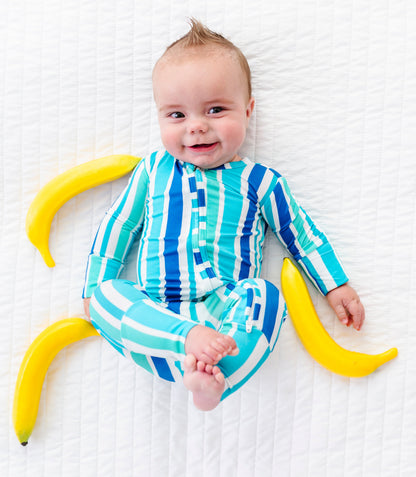 A baby lying on a white textured blanket, wearing a blue and green striped long-sleeve onesie. Two yellow bananas are placed on either side of the baby.