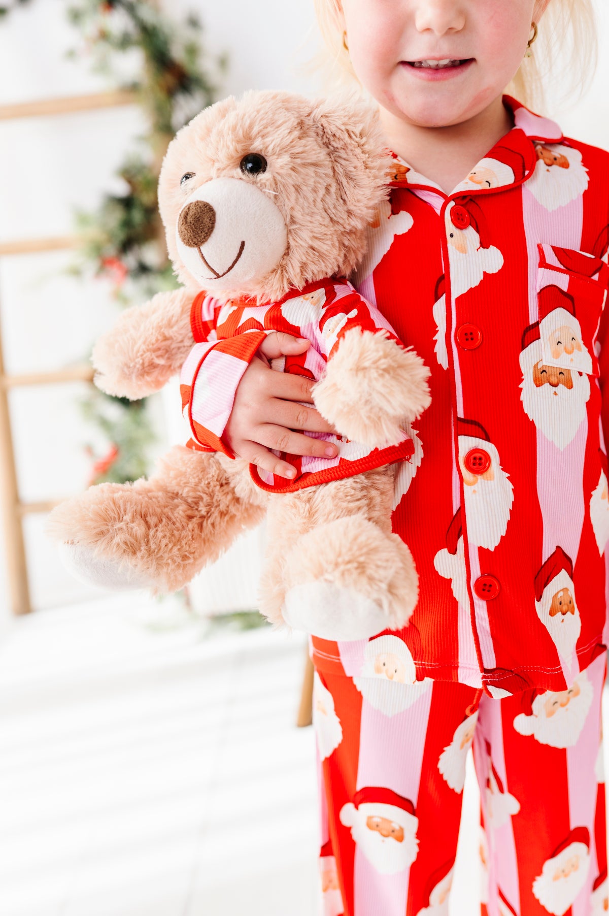 A child wearing red and pink Santa Claus bamboo pajamas holds a plush teddy bear dressed in a matching Santa outfit. The background is a softly blurred holiday setting with a Christmas tree.