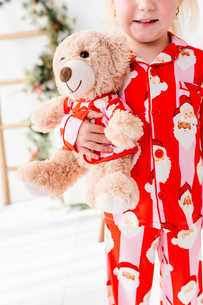 A child wearing red and pink Santa Claus bamboo pajamas holds a plush teddy bear dressed in a matching Santa outfit. The background is a softly blurred holiday setting with a Christmas tree.