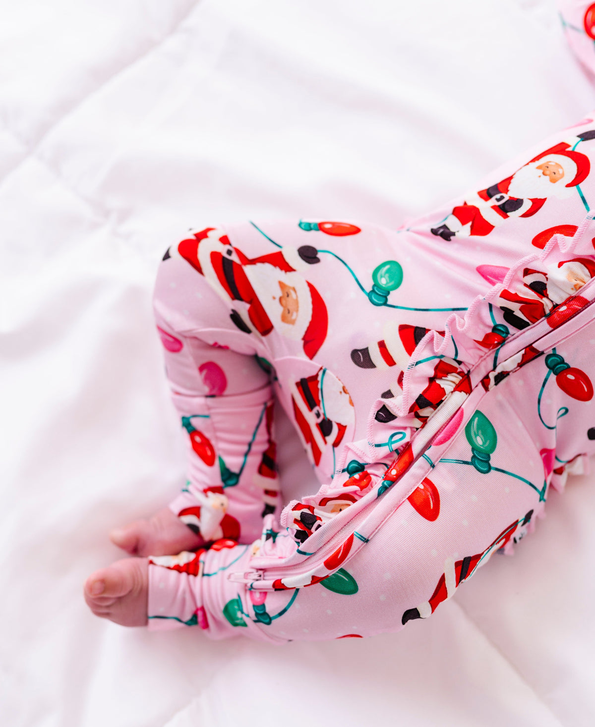 A close-up detail shot of a baby girl wearing a pink ruffle bamboo convertible romper featuring  variety of Santas hanging from multicolor Christmas lights 