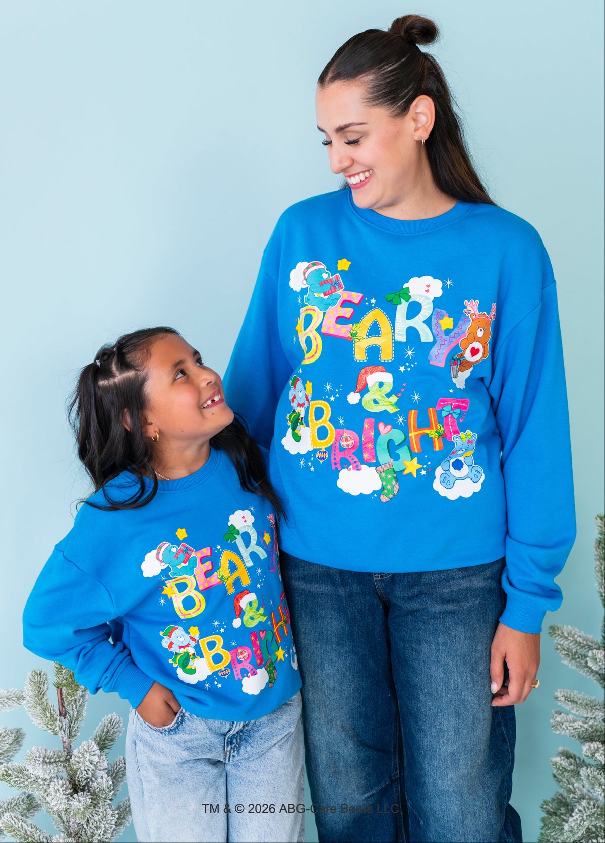 A woman and a child wearing matching bright blue crewneck sweatshirts with colorful 'Bear & Bbq Hs' holiday graphics, posed against a light blue background with small frosted Christmas trees at the sides.