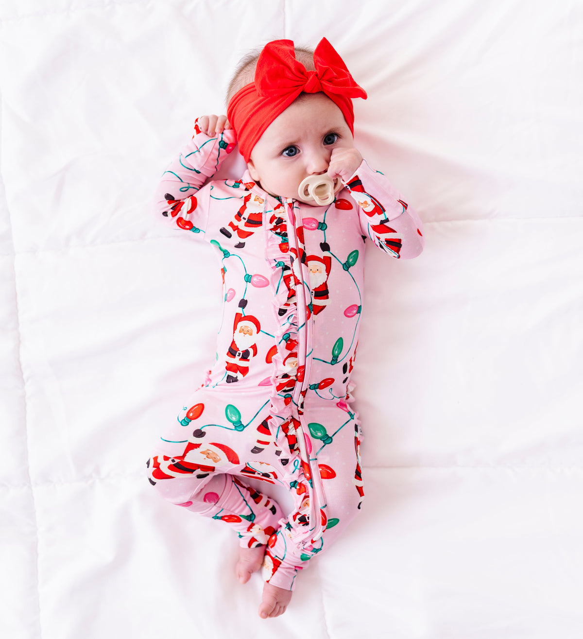 A baby girl laying on a white blanket and wearing a pink ruffle bamboo convertible romper featuring  variety of Santas hanging from multicolor Christmas lights 
