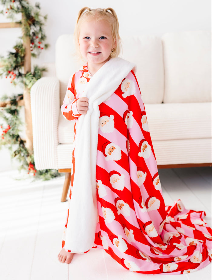 A young girl with blonde pigtails stands barefoot in a bright living room, wrapped in a red and pink Santa Claus patterned bamboo blanket with a white fleece lining.