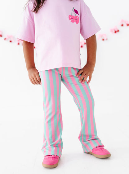 A child wearing a light pink t-shirt with a cherry graphic and pink-and-light-blue vertically striped pants, paired with pink shoes, standing against a white background.
