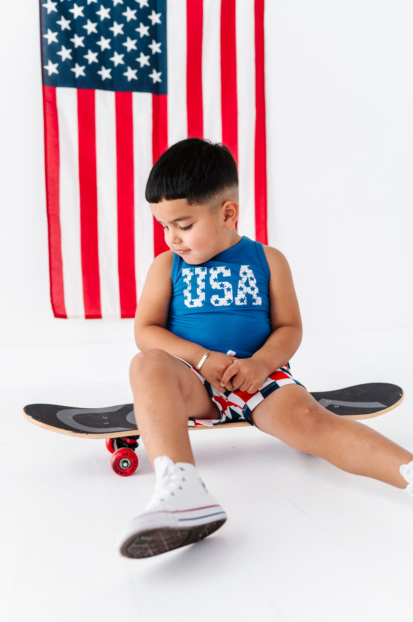 Child sitting on a skateboard wearing a bamboo-cotton "USA" tank with an American flag in the background