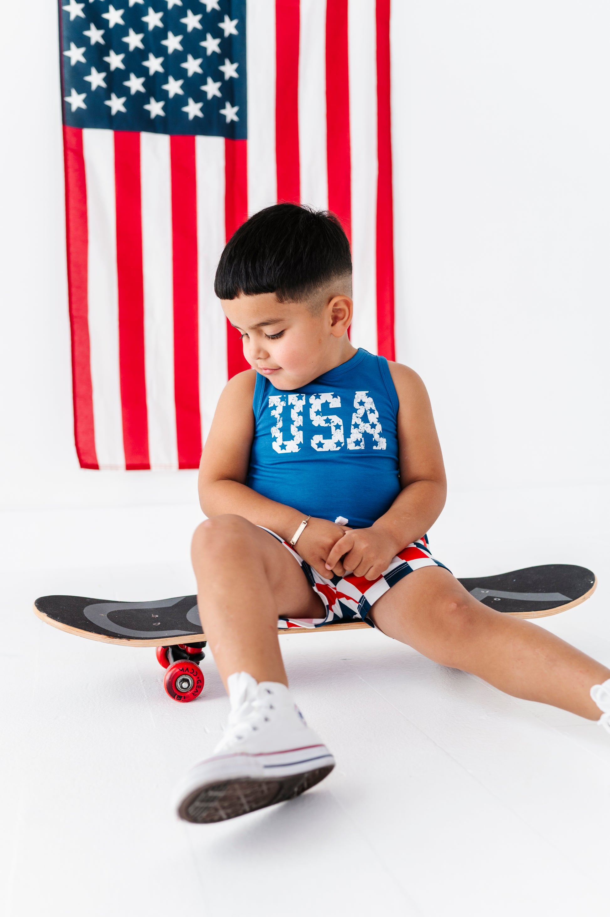 Child sitting on a skateboard wearing a bamboo-cotton "USA" tank with an American flag in the background