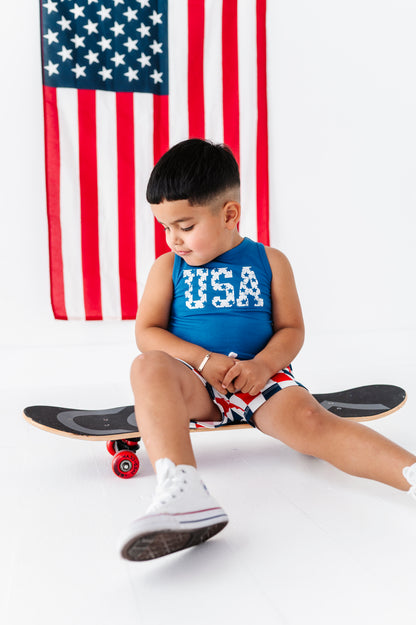 Child sitting on a skateboard wearing a bamboo-cotton "USA" tank with an American flag in the background
