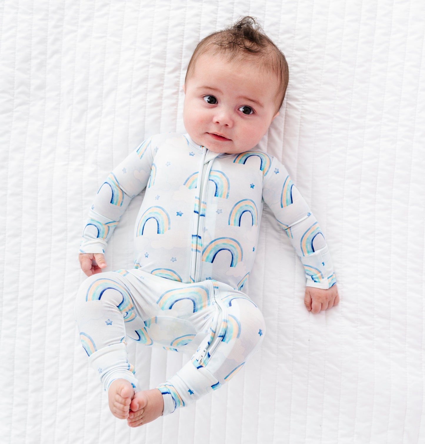 Baby lying on a white textured surface wearing a bamboo convertible romper with blue rainbow patterns.