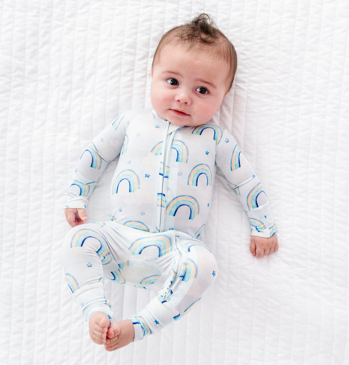 Baby lying on a white textured surface wearing a bamboo convertible romper with blue rainbow patterns.