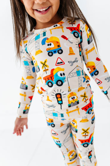 A young child wearing a long-sleeve one-piece pajama top and matching pants with a colorful construction vehicle print on a cream background, photographed against a clean white studio background.