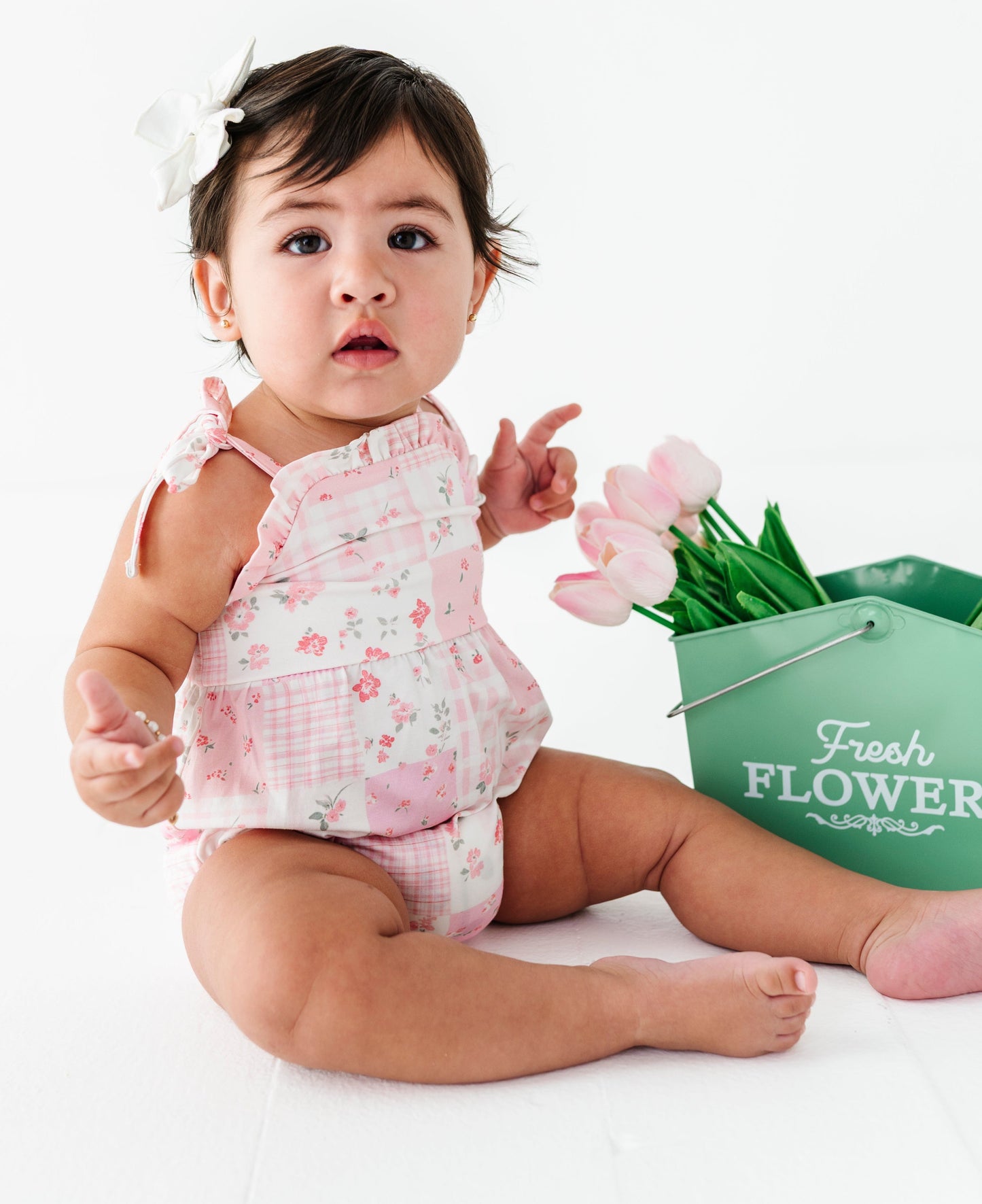 Baby in a pink floral bamboo outfit sitting next to a bucket of flowers on a white background