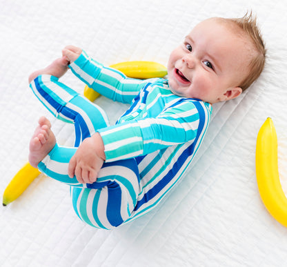 A baby lying on a white quilted blanket, wearing a blue and white striped one-piece outfit. Two yellow bananas are placed on either side of the baby.