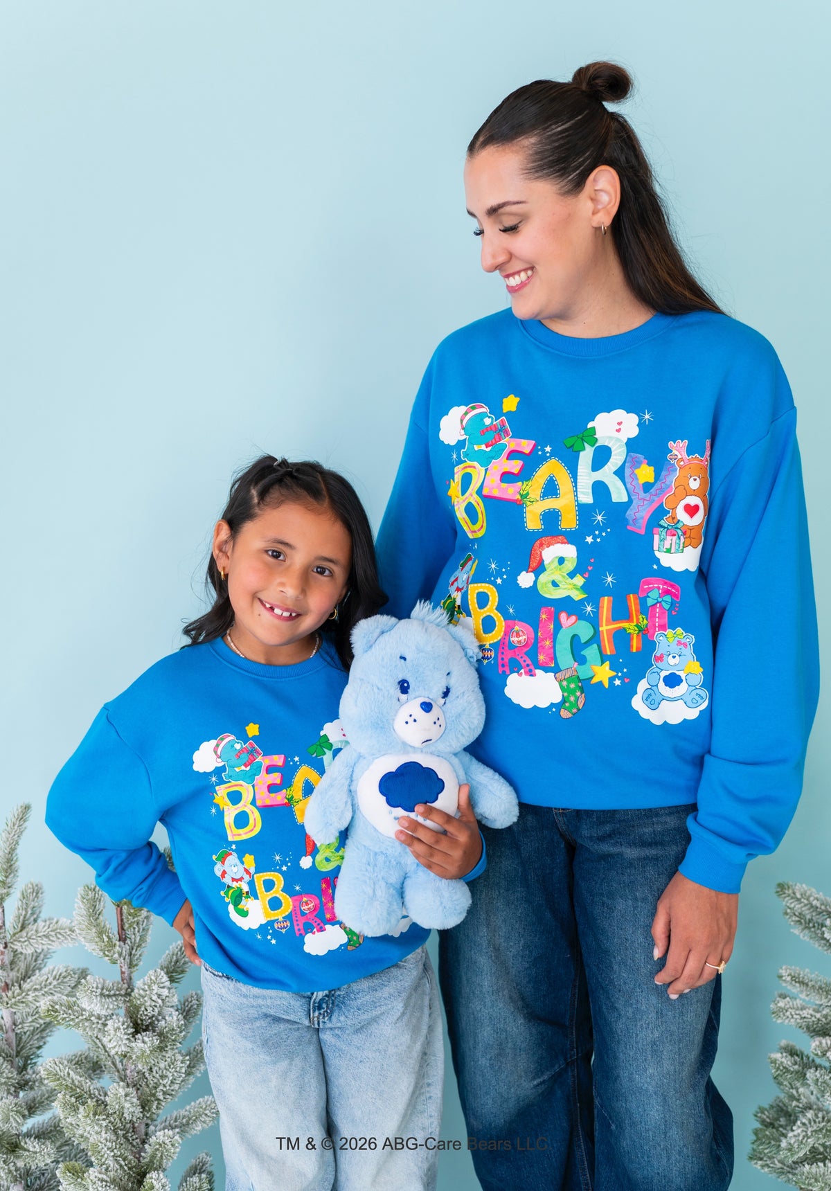 A woman and a child wearing matching bright blue sweatshirts with a colorful 'BEAR & BRIGHT' graphic, standing against a light blue background with small snowy tree decorations at the sides. The woman holds a light blue teddy bear.