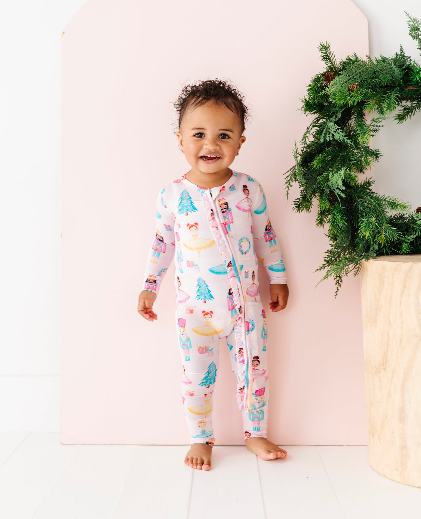Child wearing a light pink bamboo onesie with a Christmas pattern, standing next to a decorated Christmas wreath.