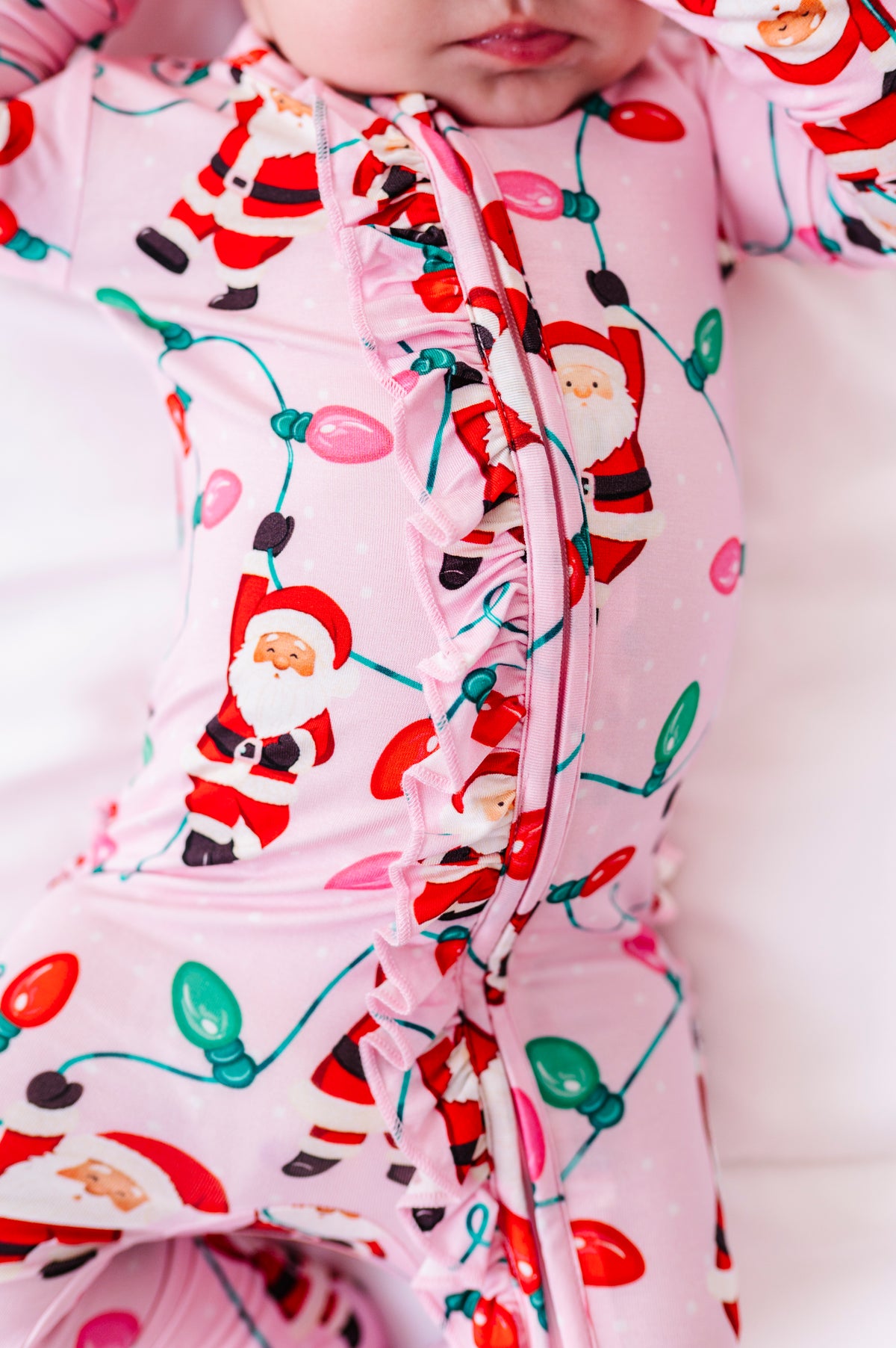 A close-up detail shot of a baby girl wearing a pink ruffle bamboo convertible romper featuring  variety of Santas hanging from multicolor Christmas lights 