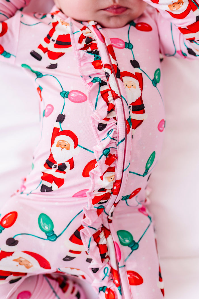 A close-up detail shot of a baby girl wearing a pink ruffle bamboo convertible romper featuring  variety of Santas hanging from multicolor Christmas lights 