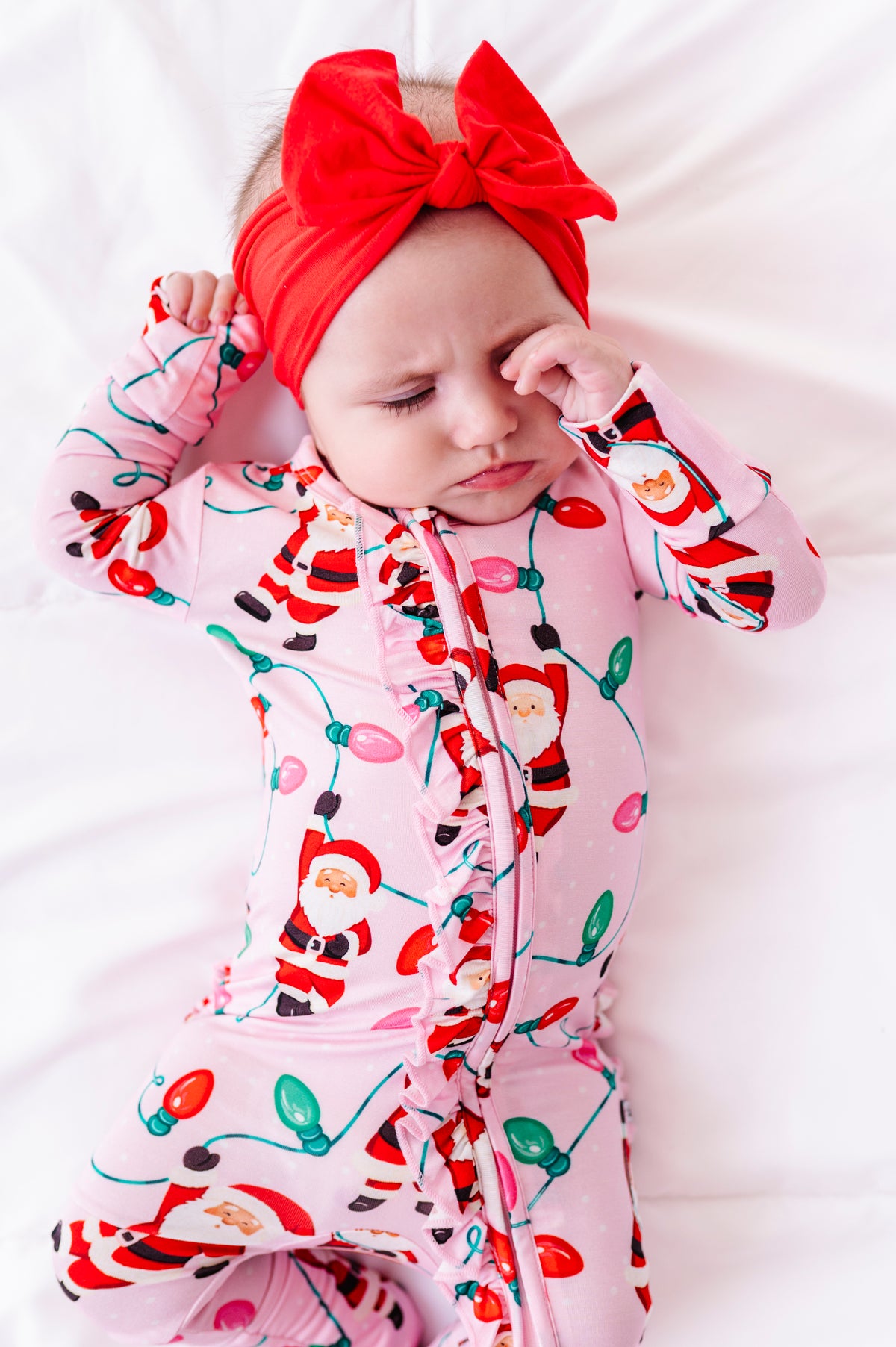 A sleepy baby girl wearing a pink ruffle bamboo convertible romper featuring  variety of Santas hanging from multicolor Christmas lights 