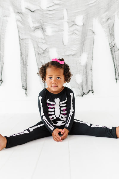 Child wearing a skeleton-themed outfit sitting on a white floor with a marble-patterned wall in the background.