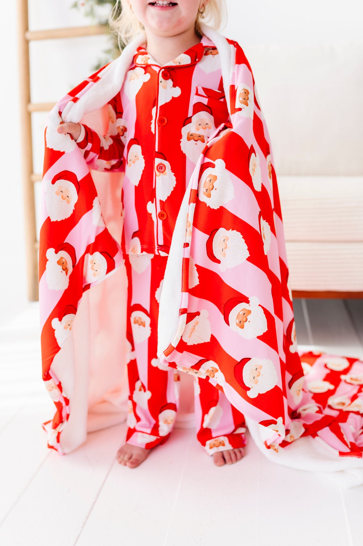 A close-up shot of a red-and-pink plush bamboo blanket. A little girl stands in a Christmas-decorated living room wearing coordinating pajamas, wrapped up in the bamboo blanket 