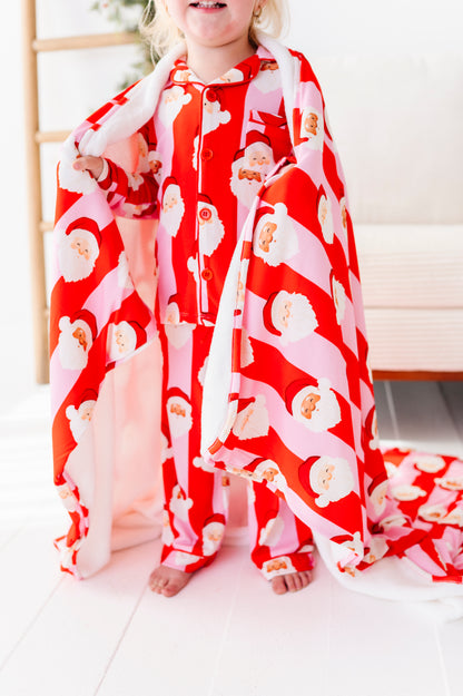 A close-up shot of a red-and-pink plush bamboo blanket. A little girl stands in a Christmas-decorated living room wearing coordinating pajamas, wrapped up in the bamboo blanket 