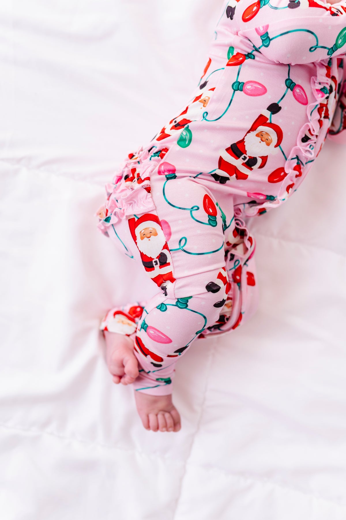 A close-up detail shot of a baby girl wearing a pink ruffle bamboo convertible romper featuring  variety of Santas hanging from multicolor Christmas lights 