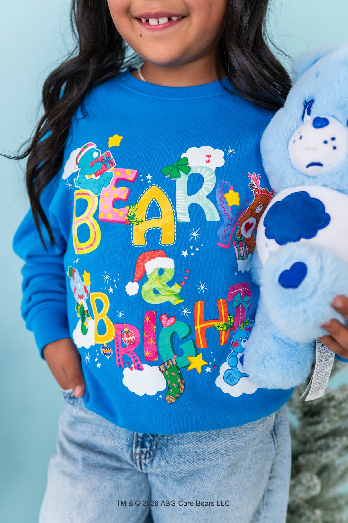 A child wearing a bright blue long-sleeve sweatshirt with a colorful 'Bear & Bright' Christmas graphic, holding a light blue Care Bear plush toy against a light blue background.