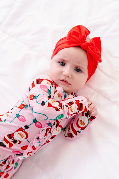 A baby girl wearing a pink ruffle bamboo convertible romper featuring  variety of Santas hanging from multicolor Christmas lights 