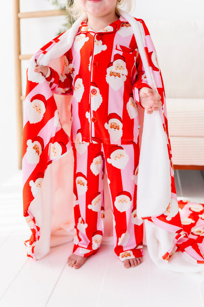 A child wearing red bamboo pajamas with a Santa Claus print, holding a matching red and pink blanket or throw in a bright indoor room.