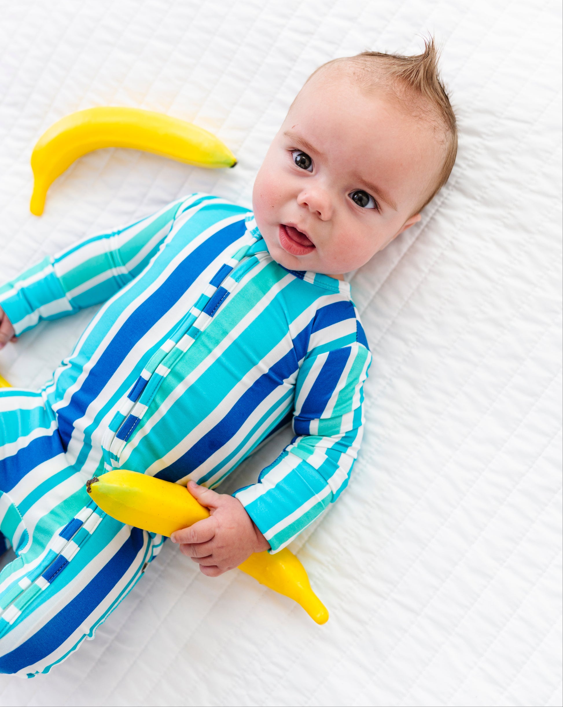 A baby lying on a white textured blanket, wearing a blue and turquoise striped one-piece outfit and holding yellow banana-shaped toys.